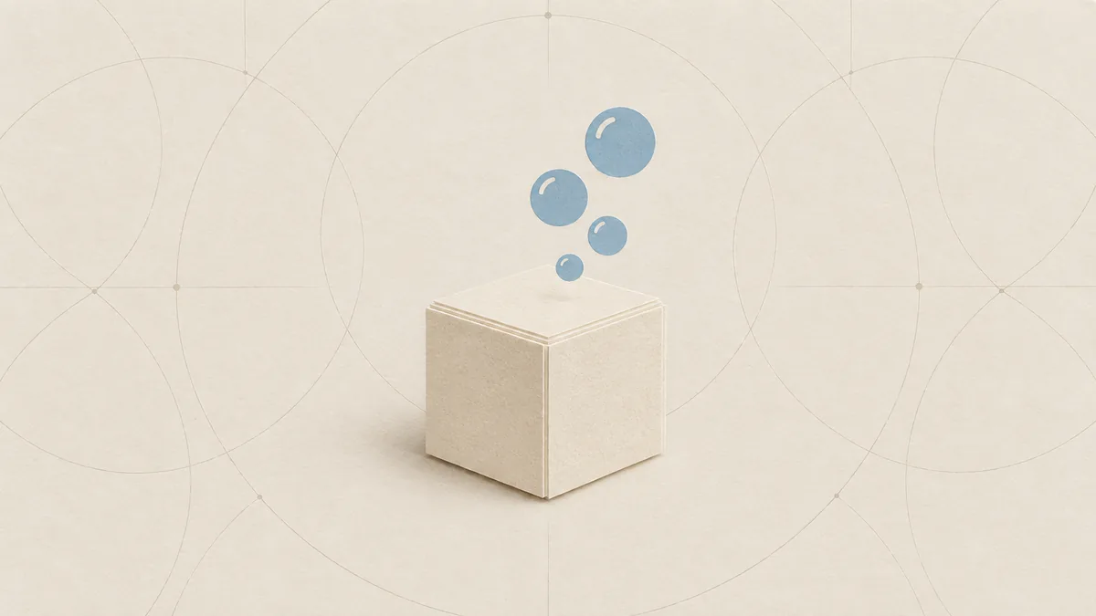 A pale cut-paper cube of soap with a few blue bubbles of different sizes rising from it, on warm off-white paper.