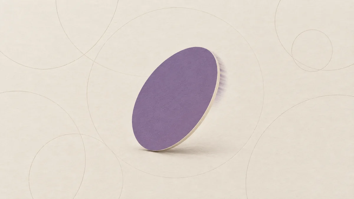 A single blank paper disc spinning on its edge, one face violet and one cream, on warm off-white paper with faint arcs.