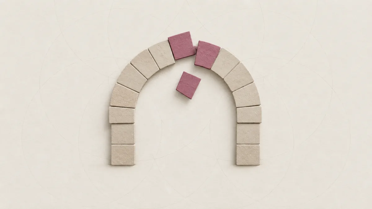 A paper stone arch, most stones set firm, with a few pink stones near its top slipping loose and one falling.